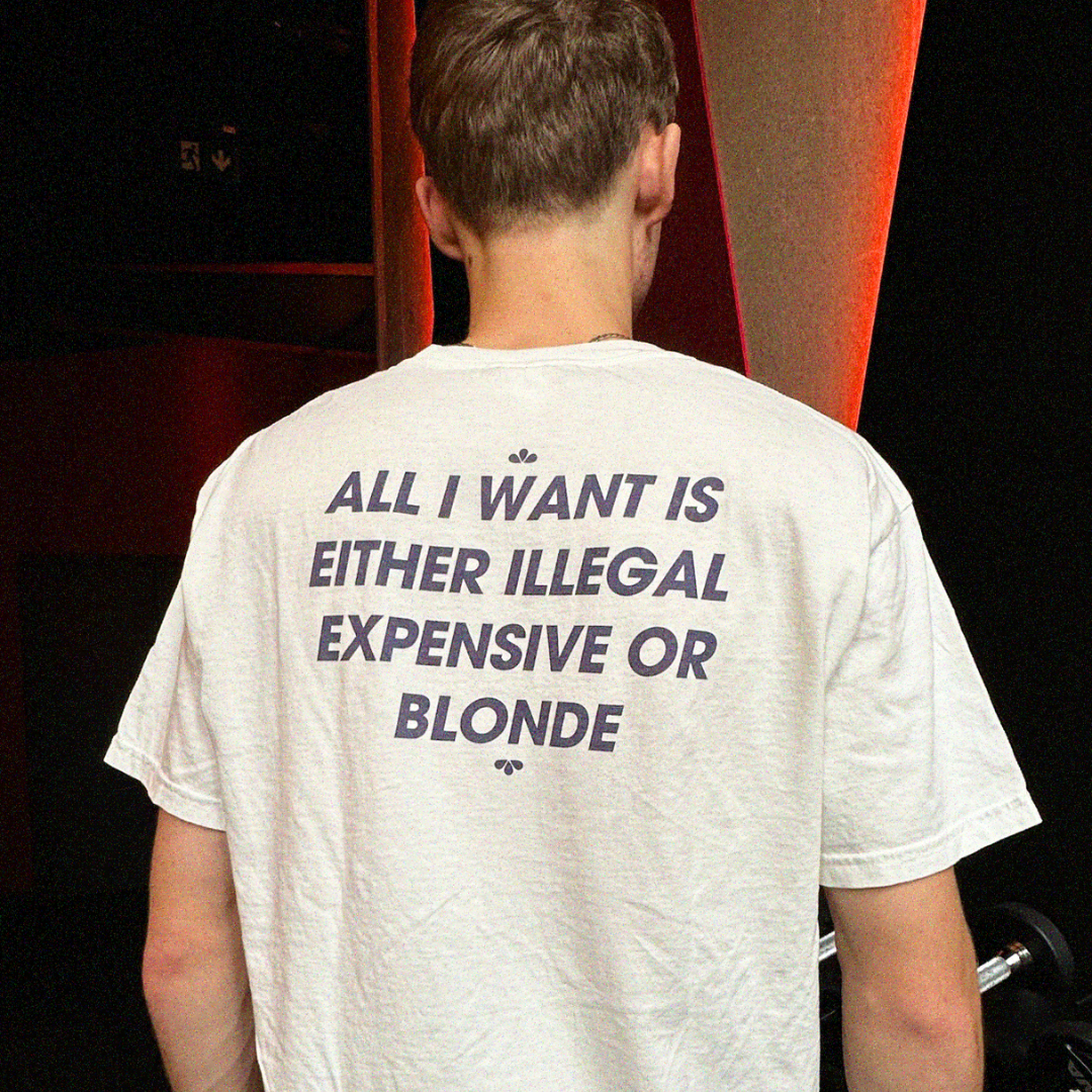 Person wearing a white T-shirt with bold black All I Want Is Illegal, Expensive or Blonde text