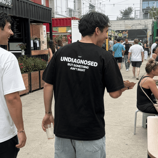 A man standing outdoors in a crowded street or market setting, wearing a black t-shirt with bold white text on the back that reads 'UNDIAGNOSED BUT SOMETHING AIN' RIGHT'.