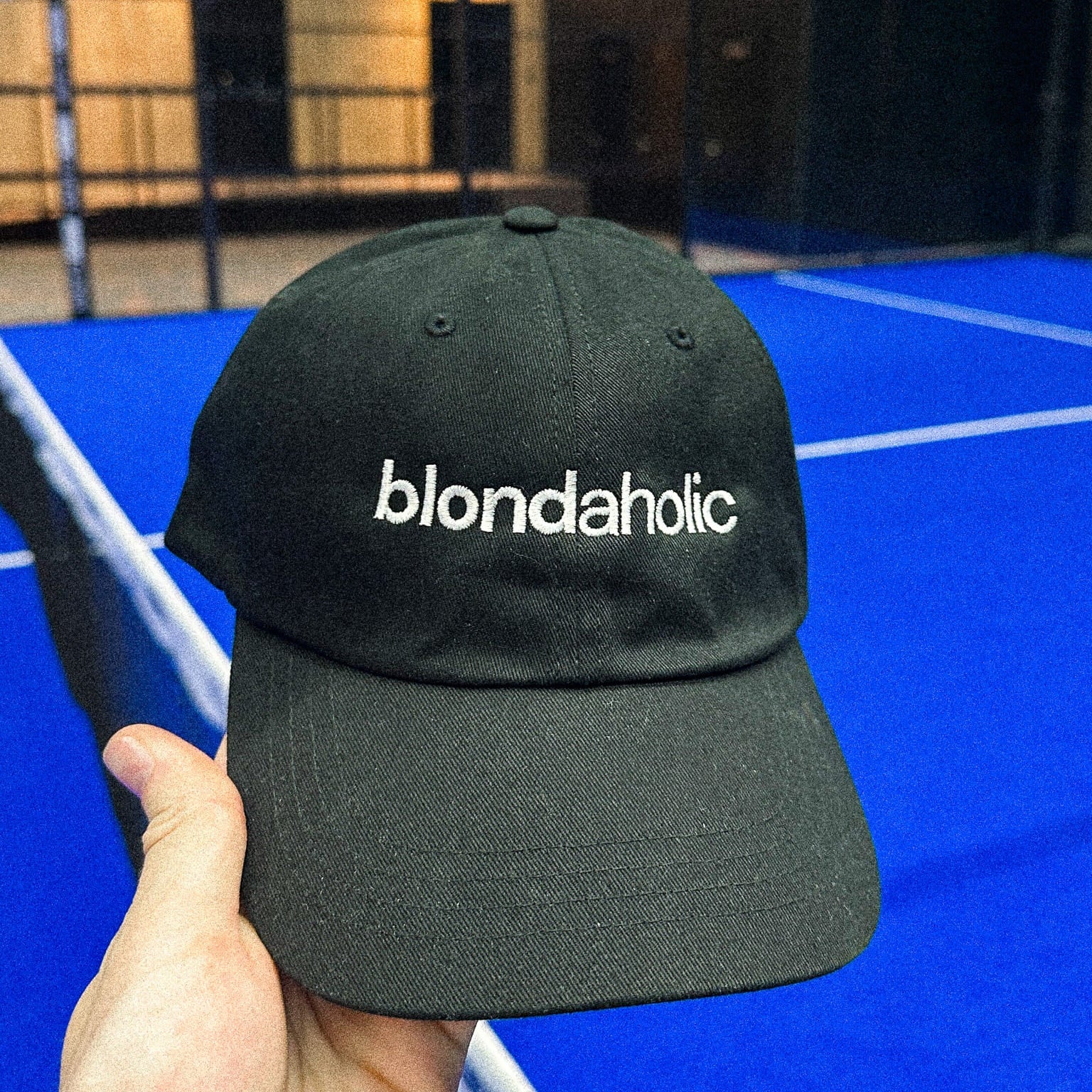 Black cap with 'blondaholic' text held in front of a blue indoor court.