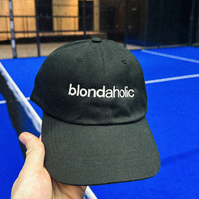 Black cap with 'blondaholic' text held in front of a blue indoor court.